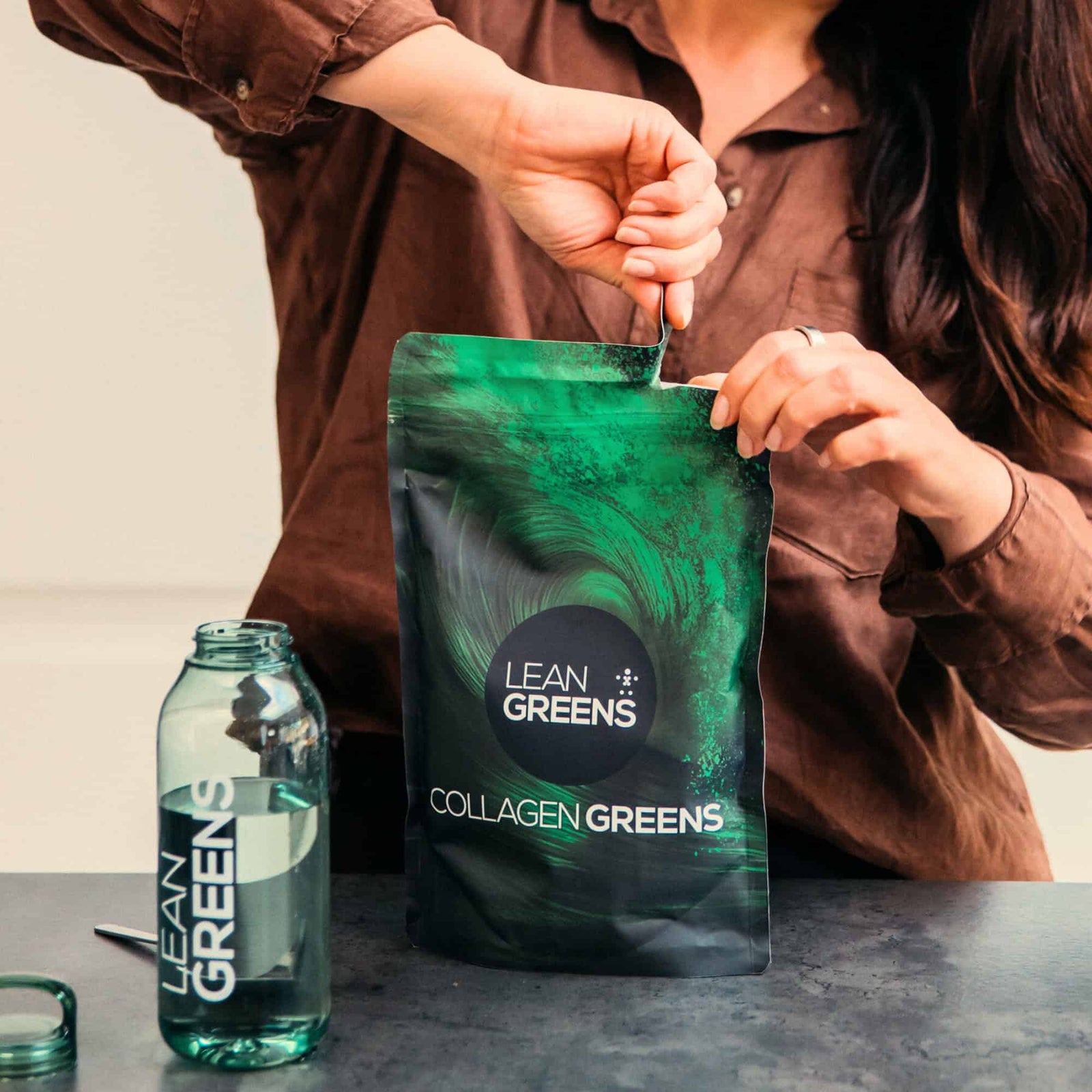 Person opening a Lean Greens Collagen Greens package with a bottle next to it on a table. UK alternative to AG1