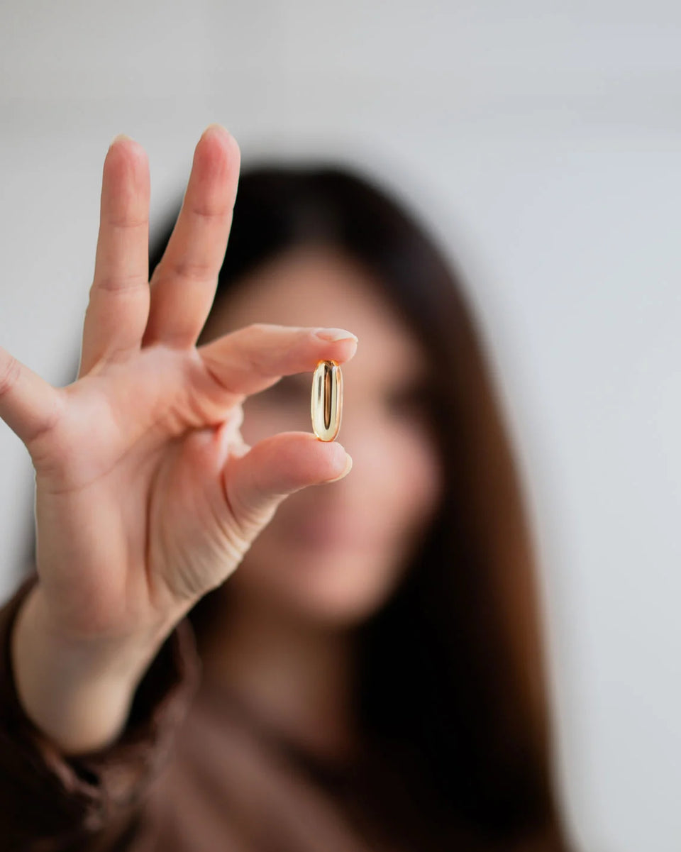 A single golden Good Fats omega-3 softgel held up between finger and thumb, the person behind it out of focus