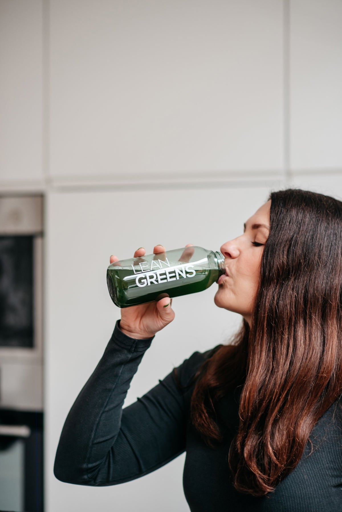 A woman drinking Lean Greens from a glass bottle at a kitchen counter
