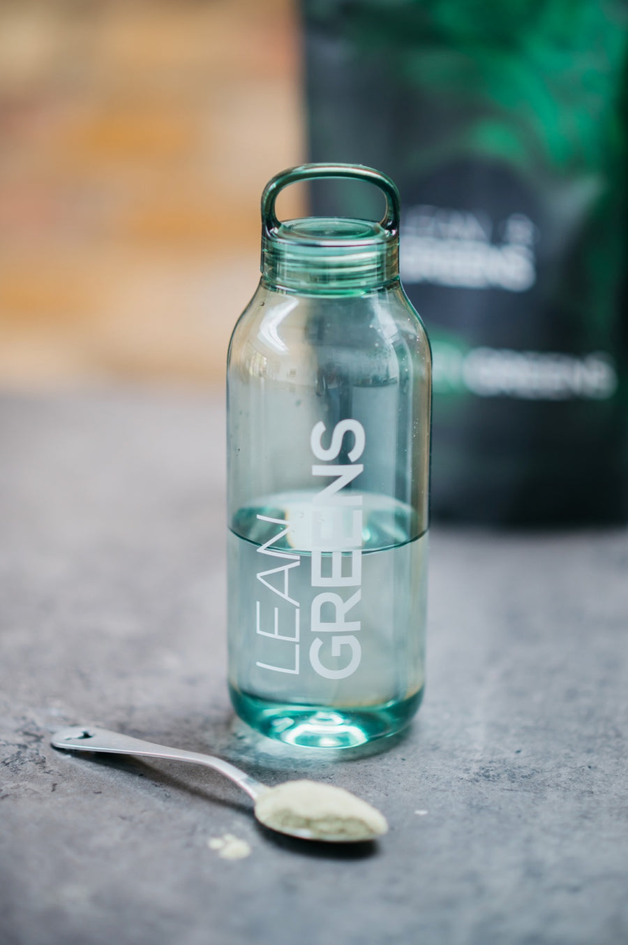 A Lean Greens glass bottle filled with cold water, a teaspoon of Collagen Greens powder beside it