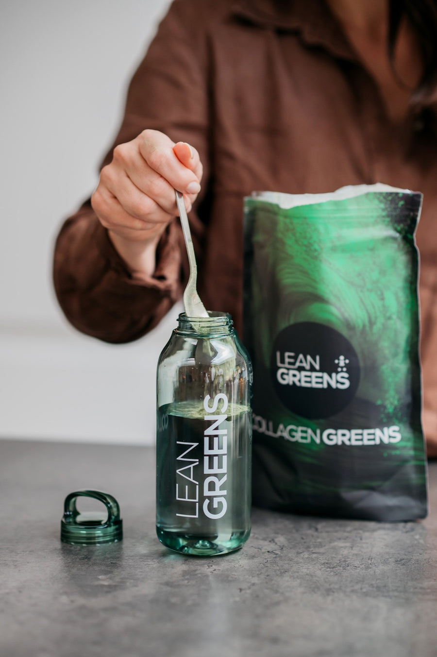 A teaspoon of Collagen Greens powder being added to a bottle of water, the open pouch beside it
